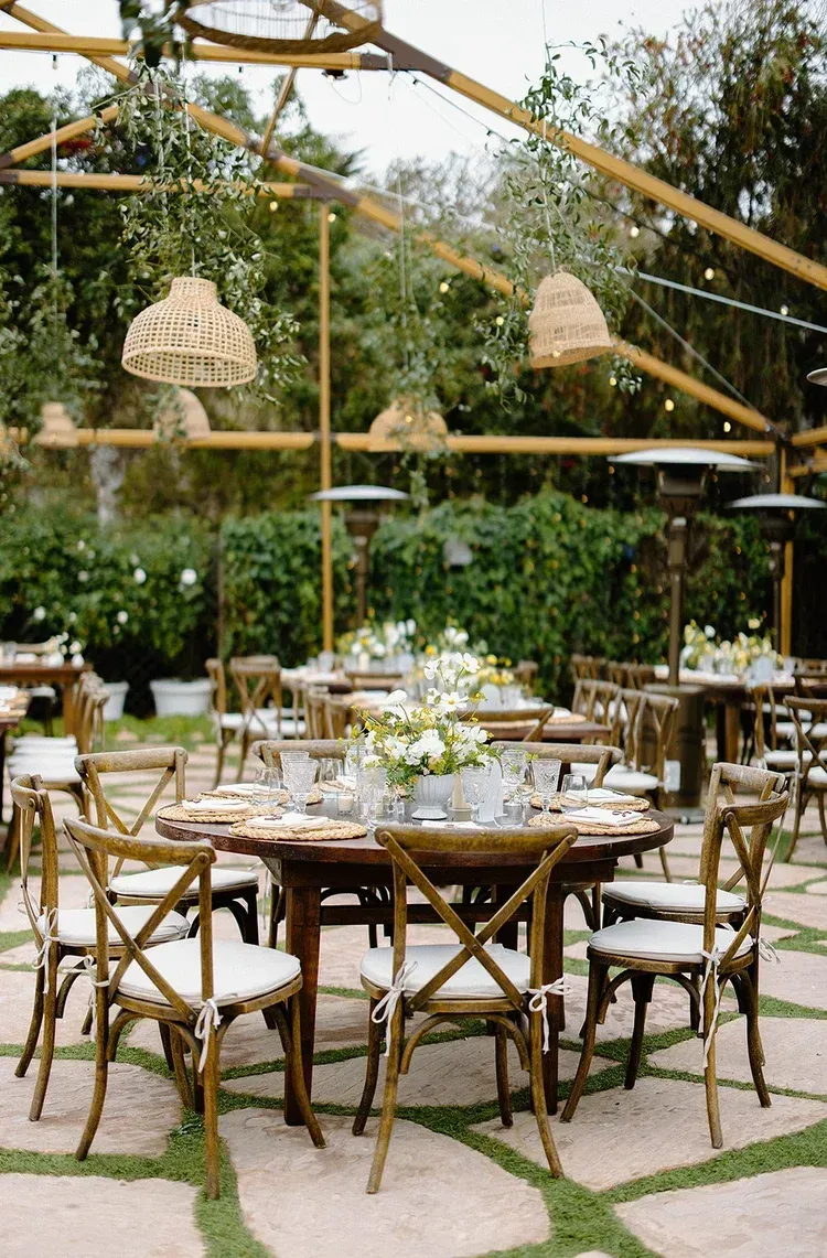 Open-air rustic banquet setup with bamboo chairs, round tables, hanging lights, and greenery.