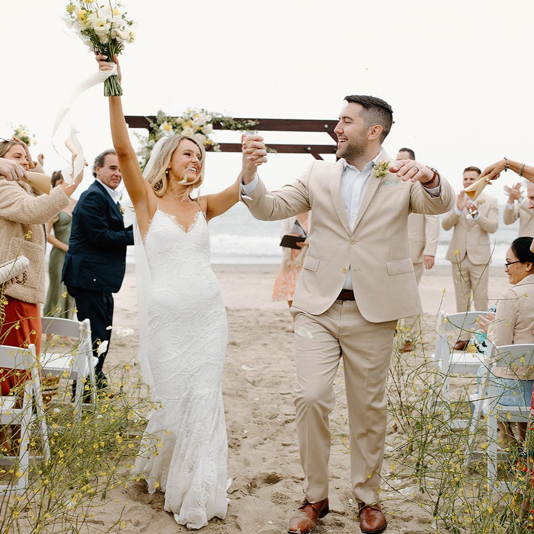 Bride and groom walk down the aisle outdoors, smiling and holding flowers with guests in the background