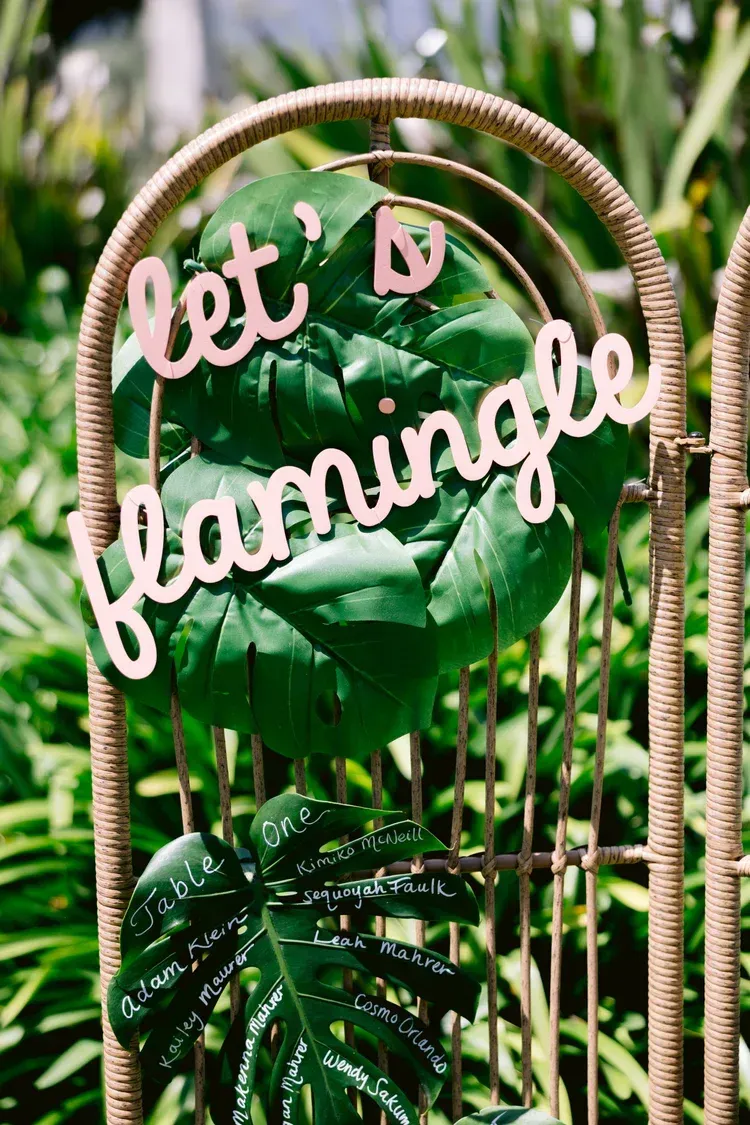 Pink cursive “let’s flamingle” sign on a woven arch amid lush green tropical leaves