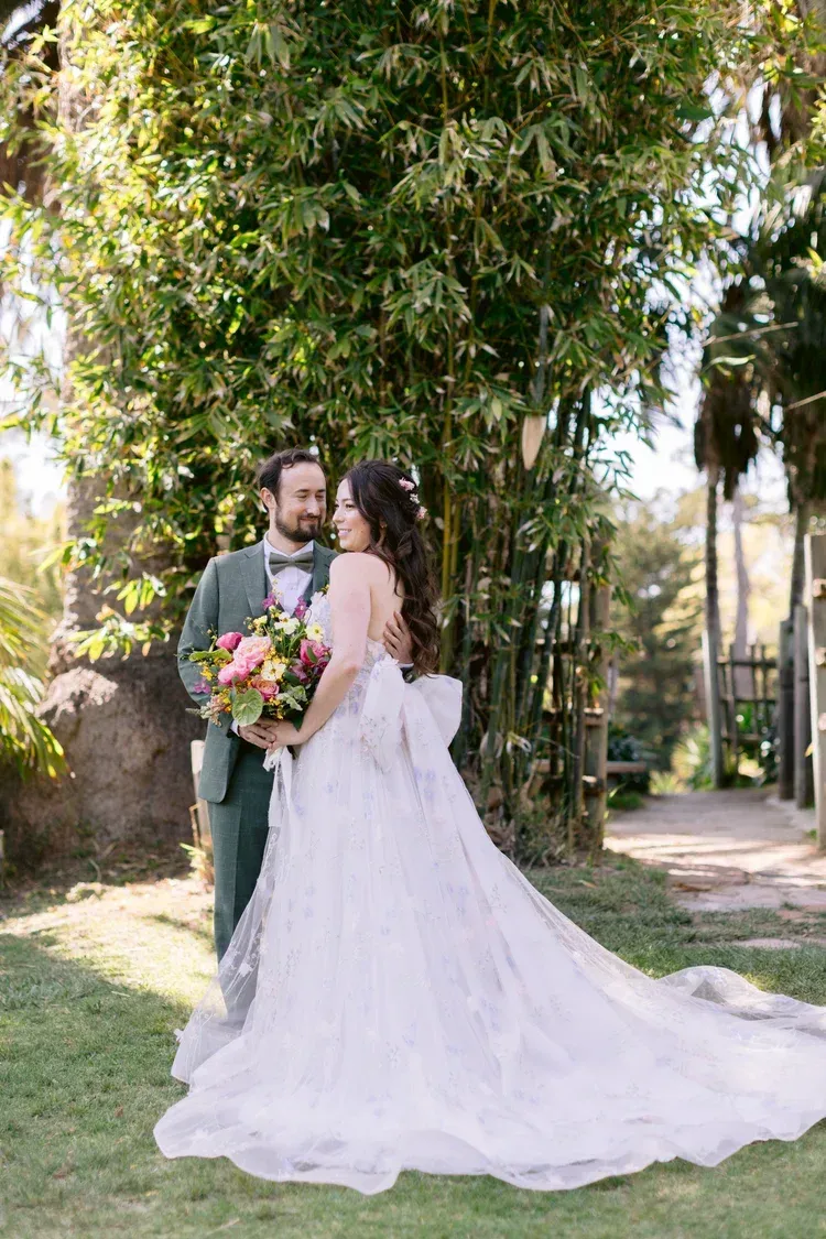 Bride and groom in a garden under a tree, holding a bouquet and facing each other in wedding attire