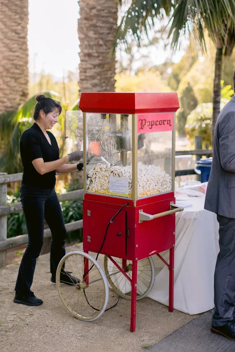 Person operating a red popcorn cart outdoors near palm trees
