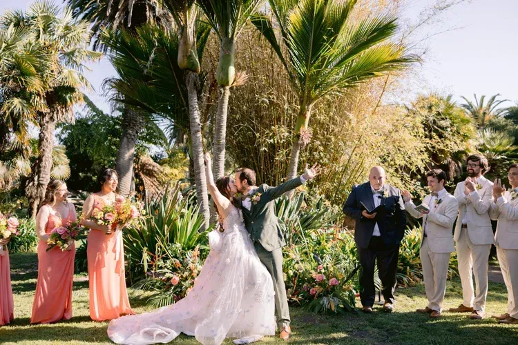 Wedding couple dancing in a sunny garden, surrounded by attendants and guests with floral arrangements.