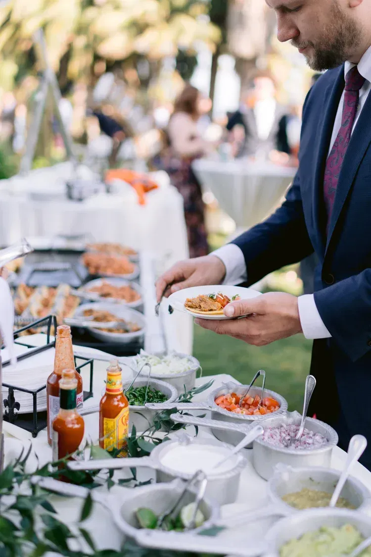 Man in a suit serving food at an outdoor buffet table with bottles and dishes