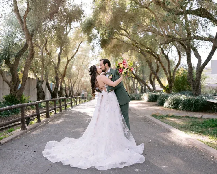 Bride and groom embracing on a tree-lined path, holding a colorful bouquet and wearing a white gown and dark suit.