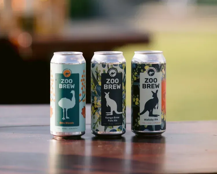 Three Zoo Brew cans on a wooden table, with white bird silhouettes and blurred background.