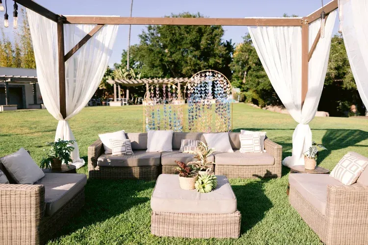 Outdoor lounge with wicker seating, white curtains, and a wooden pergola on a sunny lawn