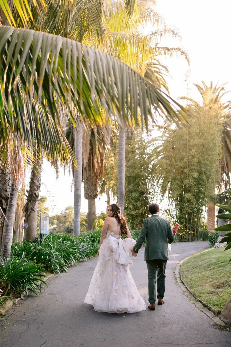 Bride and groom walking hand in hand along a palm-lined garden path