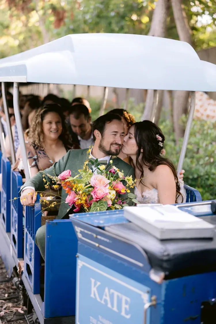 Couple riding in a blue wedding trolley under a white canopy, surrounded by flowers and smiling guests