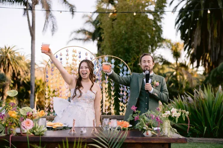 Couple at outdoor wedding reception, bride raising arm beside groom holding microphone, with floral décor.