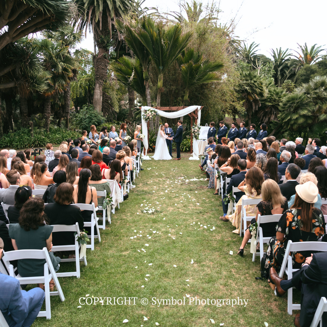Outdoor wedding ceremony in a garden, with guests seated on white chairs facing the couple under a floral arch.