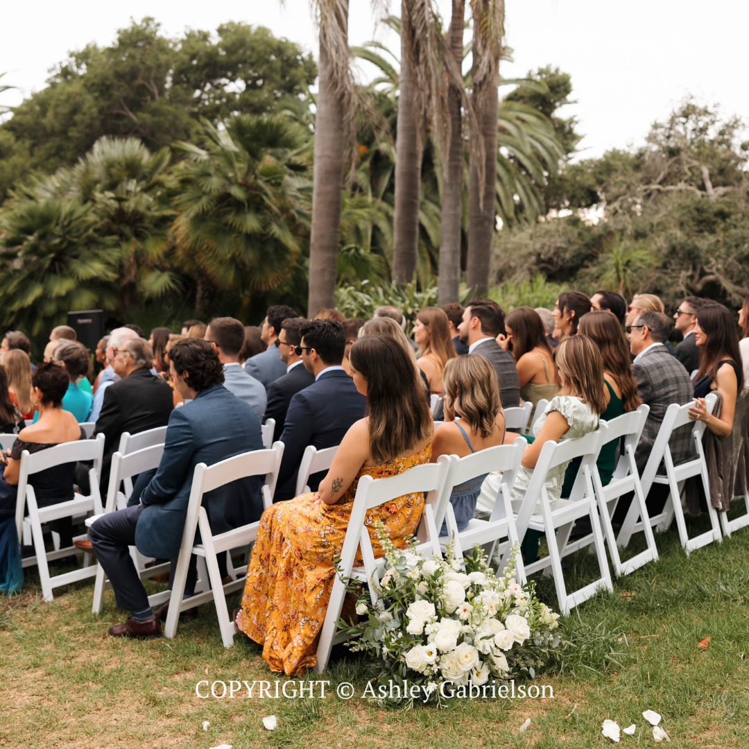 Guests seated outdoors on white chairs at a garden ceremony, with palm trees and floral arrangements nearby