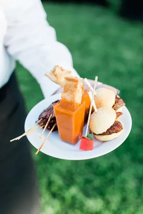 A hand holding a white plate with assorted desserts, including a small orange drink, outdoors on grass.