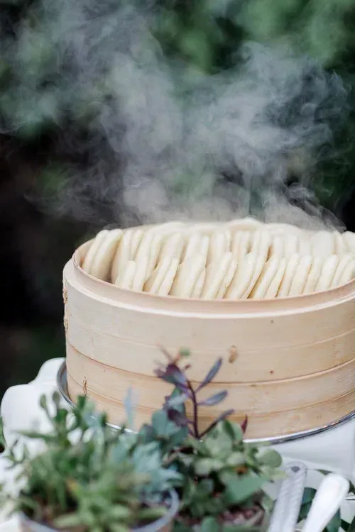 Steam rises from a bamboo steamer filled with dumplings, with herbs in the foreground.