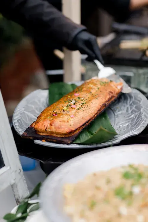 Grilled salmon fillet on a banana leaf, being served with tongs at a food stall