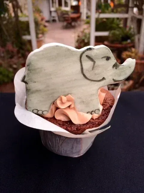 Elephant-shaped cookie on a cupcake in a white wrapper, outdoors with a blurred garden background