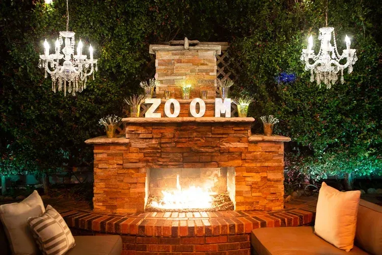 Outdoor lounge with a brick fireplace, glowing fire, ZOOM sign, and chandeliers at night