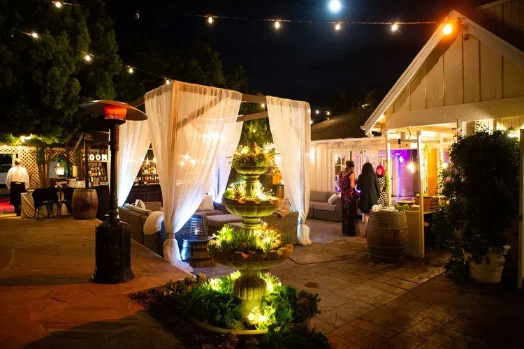 Nighttime outdoor wedding reception with lit fountain, white drapes, string lights, and guests near a decorated pavilion