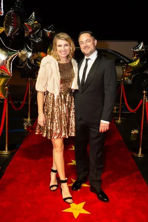 A couple posing on a red carpet with balloons and stanchions in the background.