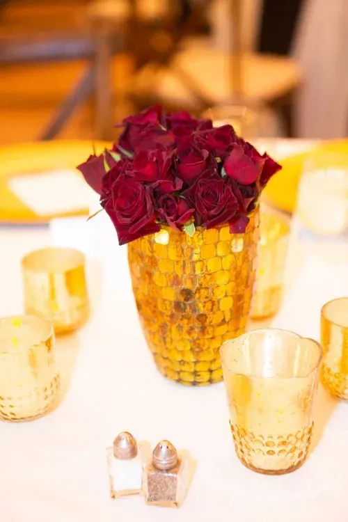 Red roses in a gold vase on a white table, surrounded by gold votive candles.