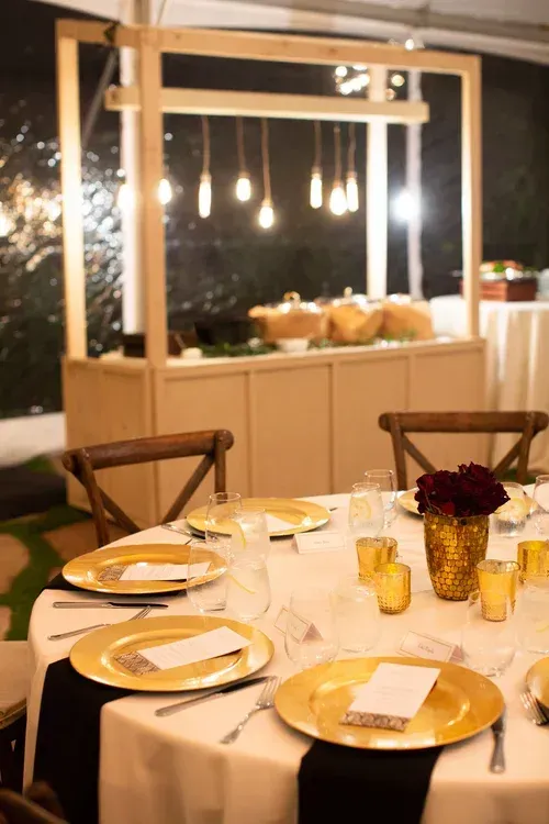 Set dining table with white tablecloth, gold plates, and candlelit buffet in the background