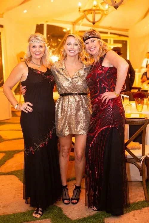 Three women posing together at a warmly lit indoor event, wearing black and gold dresses.