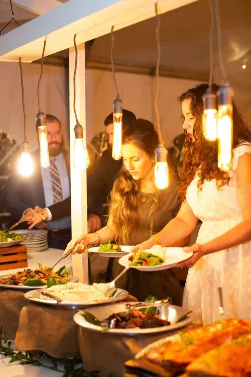 People serving food at a warmly lit buffet table under hanging lights.