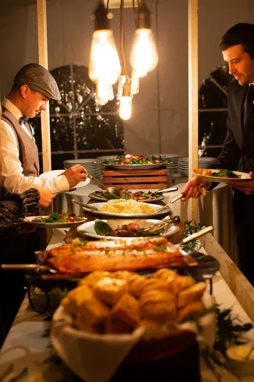 Two people serving food at a warmly lit buffet table with assorted dishes and candles