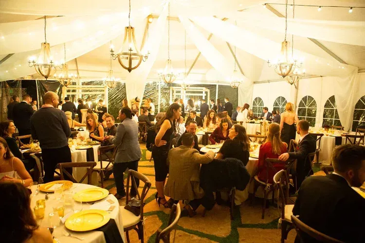 Guests mingling under a white tent at a warmly lit indoor-outdoor event with tables and chandeliers