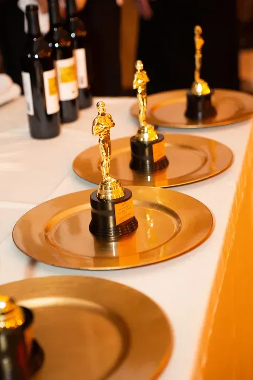 Gold award statuettes on copper plates at a ceremony table, with wine bottles in the background
