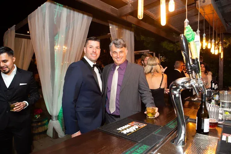Two men posing at a bar, with drinks and warm hanging lights in the background.