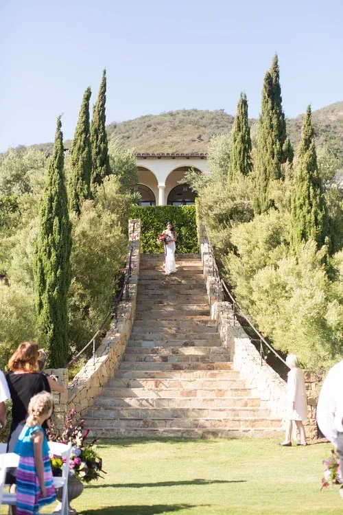 Stone stairway through cypress trees to a hillside archway, with people walking up in a garden.