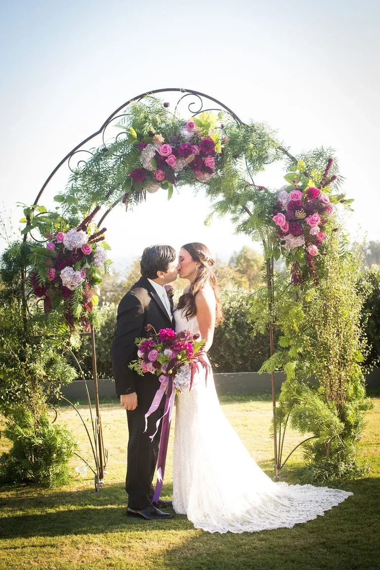 Bride and groom kissing beneath a flower-covered arch outdoors