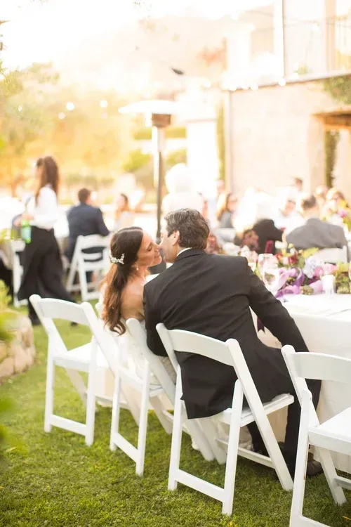Guests seated at white chairs for an outdoor wedding reception, with floral tables in soft sunlight
