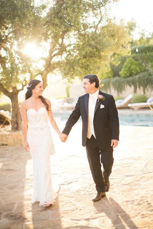 Couple in formal attire holding hands and walking outdoors by a pool at sunset.