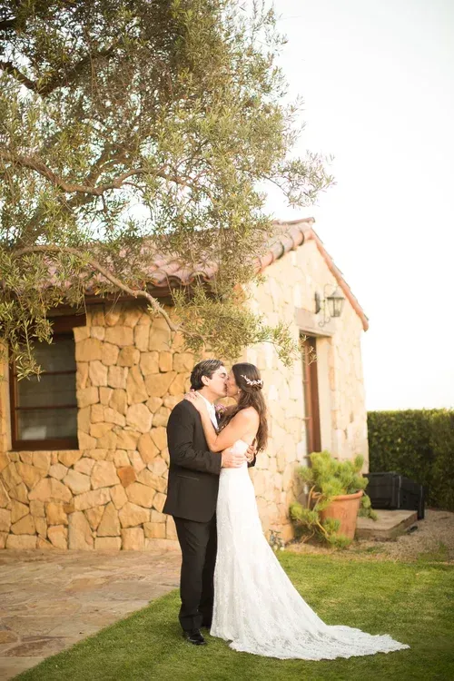 Bride and groom kissing beside a rustic stone house in a garden