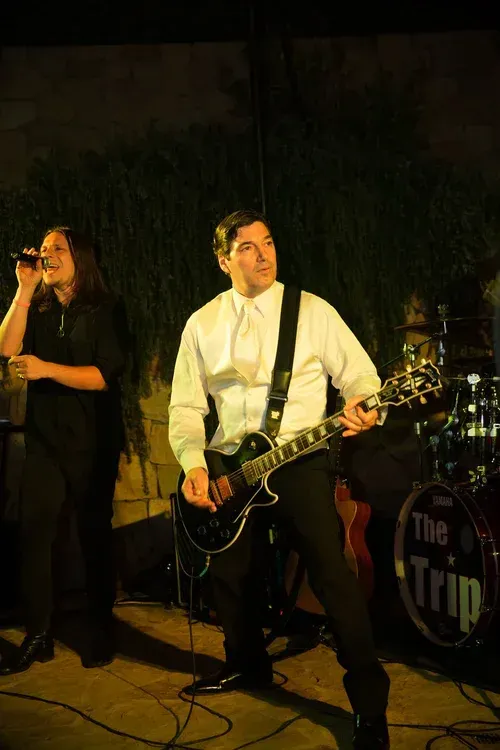 Live band performing on a dimly lit stage, with a singer at left and guitarist at center.