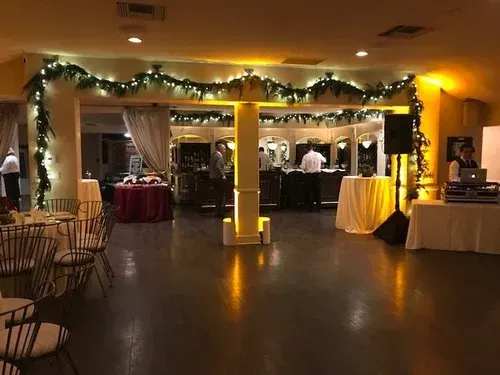 Decorated banquet hall with holiday garlands, round tables, and a lit dessert table at the back
