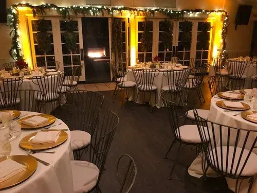 Empty banquet room with round tables, white chairs, and warm string lights by large windows