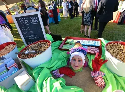 Person peeking from a popcorn bin at an outdoor fair with colorful treats and a chalkboard sign