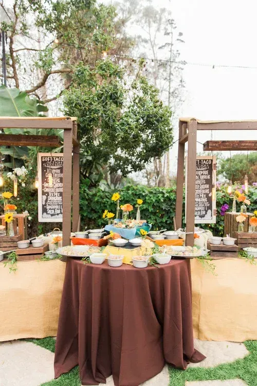 Dessert buffet table with brown cloth, fruit and sweets, set outdoors among floral decorations and wooden frames