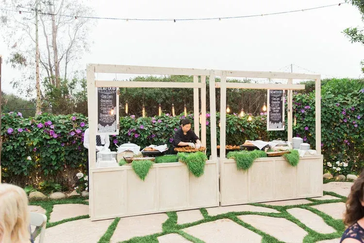 Outdoor market stall with green foliage decor and two vendors serving food under a white canopy