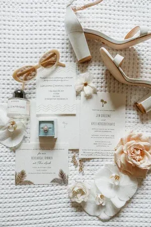 Bridal flat lay with invitation cards, beige heels, sunglasses, flowers, and jewelry on a white knit background