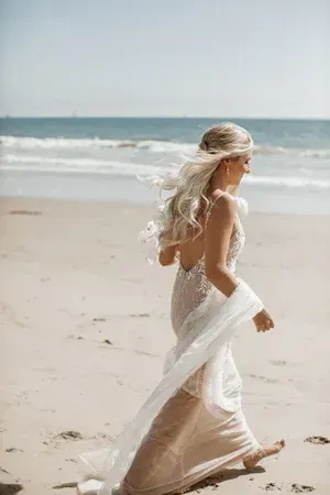 Woman in a flowing white dress walking barefoot along a sandy beach by the ocean