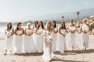 A group of women in white dresses holding bouquets on a beach with mountains in the background