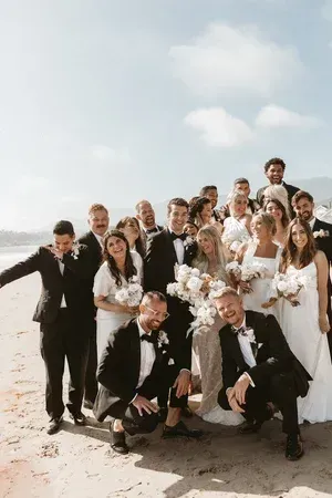Wedding party posing on a beach, smiling in white formalwear against a bright sky.