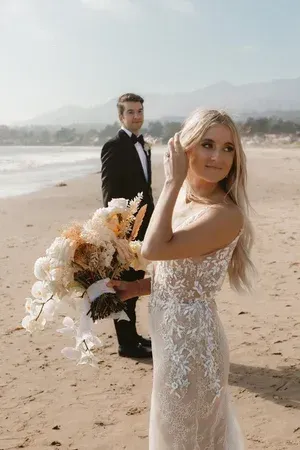 Bride holding bouquet on a beach, with groom standing behind her near the shoreline.