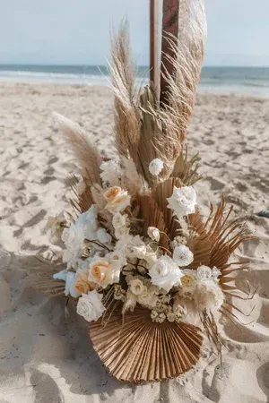 Beachy floral arrangement in neutral tones with shells and pampas grass on sand, ocean in background