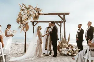 Beach wedding ceremony under a wooden arch with floral arrangements, bride and groom exchanging vows, guests nearby