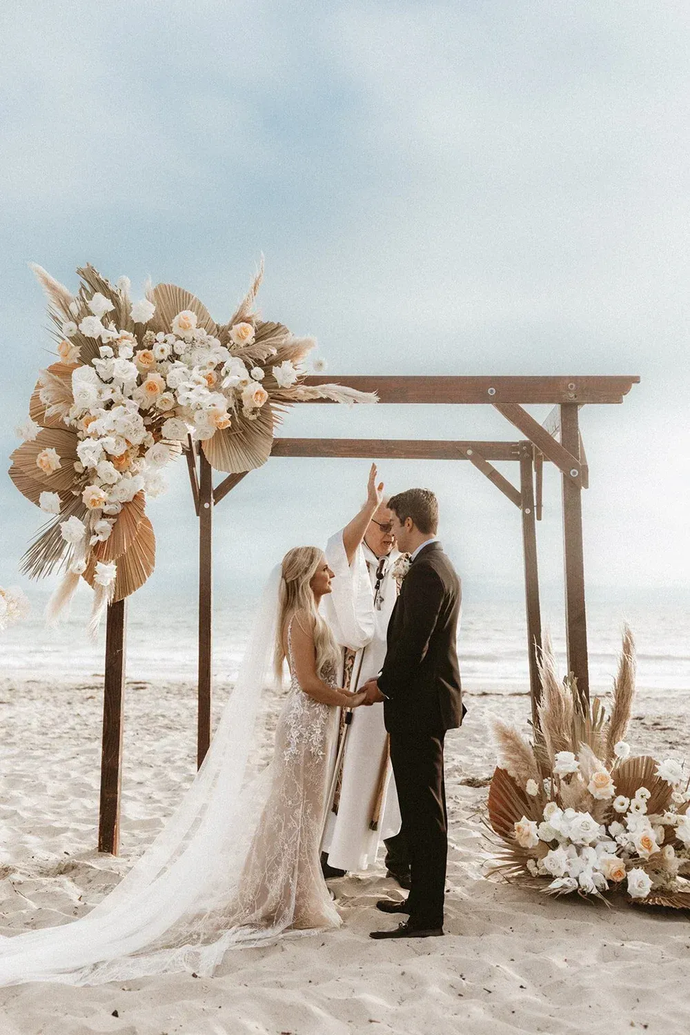 Bride and groom kissing under a wooden arch on a beach, surrounded by white flowers