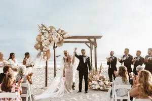 Beach wedding ceremony with couple under floral arch and guests applauding by the water